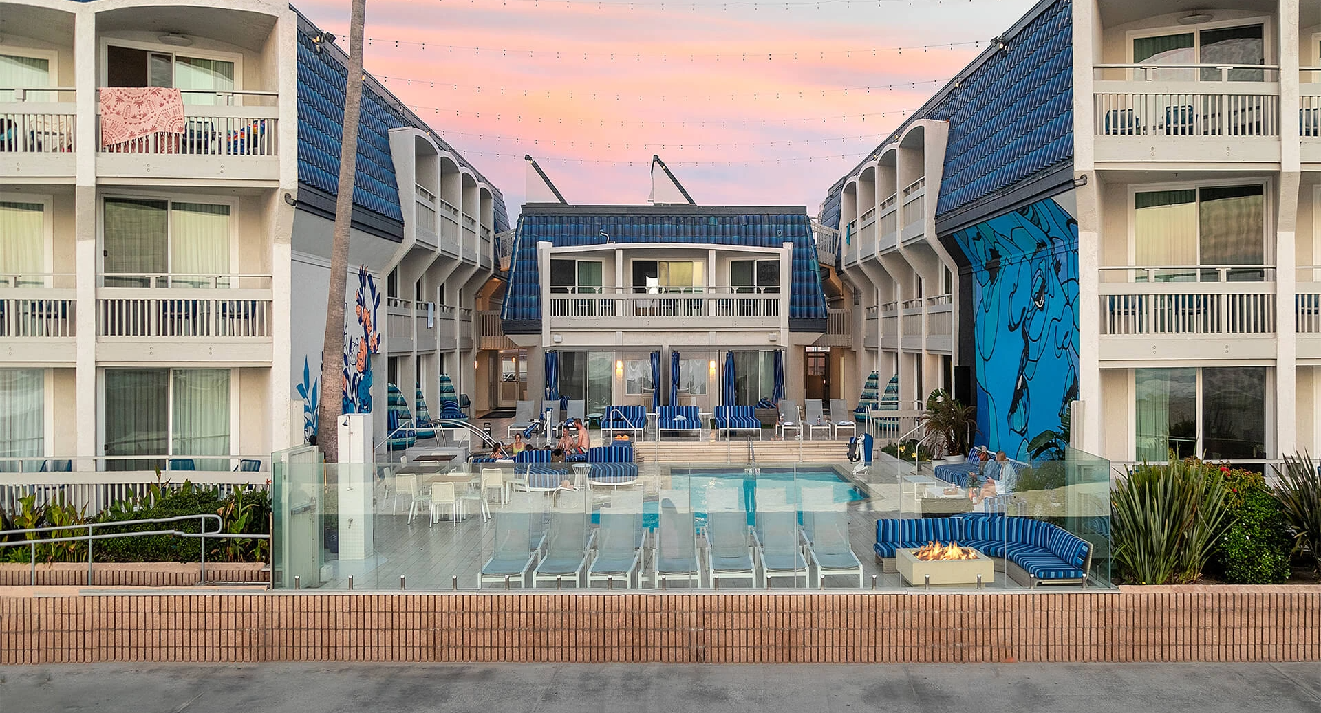 A luxury resort pool area at sunset, with blue-tiled buildings, loungers, glass railings, palm decor, and pastel sky behind the courtyard. ending with period.
