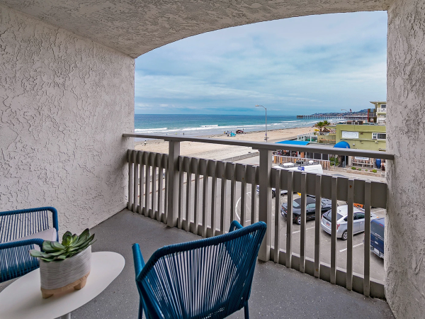 A balcony with blue chairs and a small plant, overlooking a beach and town with cars below, calm sea and cloudy sky, inviting and relaxing.