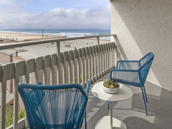 Two blue wicker chairs and a small round table on a seaside balcony, overlooking a sunny beach and ocean. End.