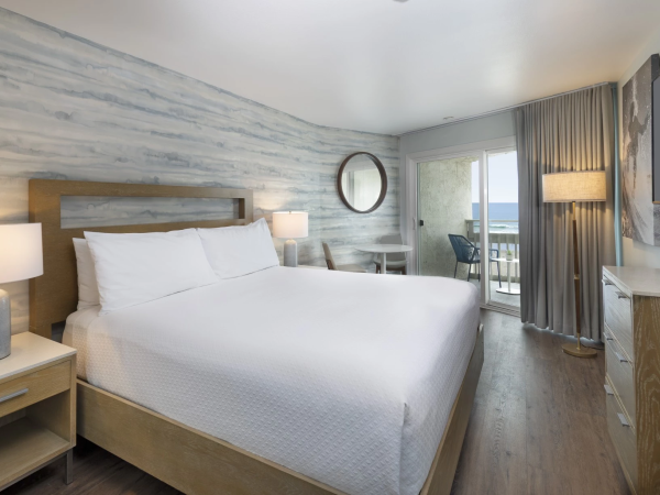 A modern hotel room with a king bed, soft white bedding, wood furniture, a round mirror, and a balcony view through glass doors.