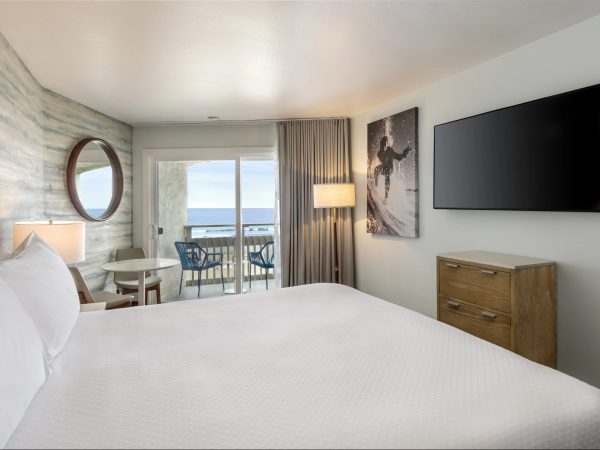 A hotel room with a large white bed, a wall-mounted TV, wooden dresser, round mirror, balcony with ocean view, and soft lighting&mdash;cozy and modern.