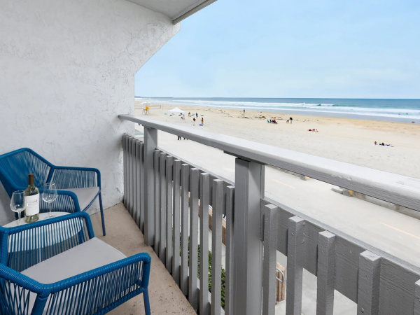 A balcony with two blue chairs and a small table holding drinks, overlooking a sandy beach and the ocean under a clear sky.