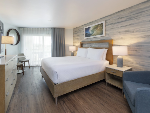 A modern hotel room with a king bed, wooden accents, blue-gray walls, two nightstands, a sofa, desk, and a large window letting in light.