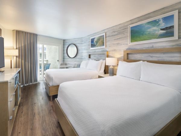 A bright hotel room with two white beds, wooden headboards, a desk, flat-screen TV, and a large window with curtains by the sea view.