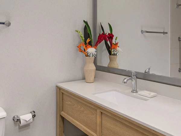 A modern bathroom vanity with a large mirror, wooden cabinet, and a light over the sink; colorful flowers in a vase sit on the counter, with towels neatly stored below.