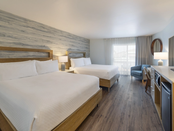 Two white‑bedroom hotel rooms with two double beds, a desk, TV, small seating area, and a window with sheer curtains.