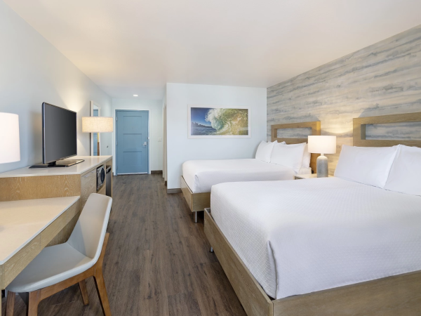 A bright hotel room with two queen beds, a desk and chair, a wall-mounted TV, bedside lamps, and a blue door at the far end, clean and airy.