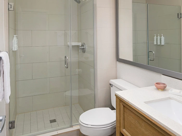 A compact bathroom with a glass-enclosed shower, wall-mounted toilet, wooden vanity with a white countertop, a large mirror, and neutral tile flooring.