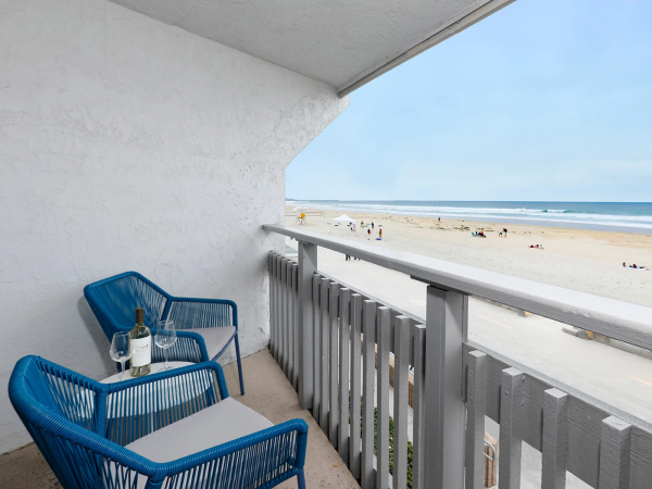 A balcony with two blue wicker chairs and a small table overlooking a sunny beach; the ocean is calm with people sunbathing near the shore.