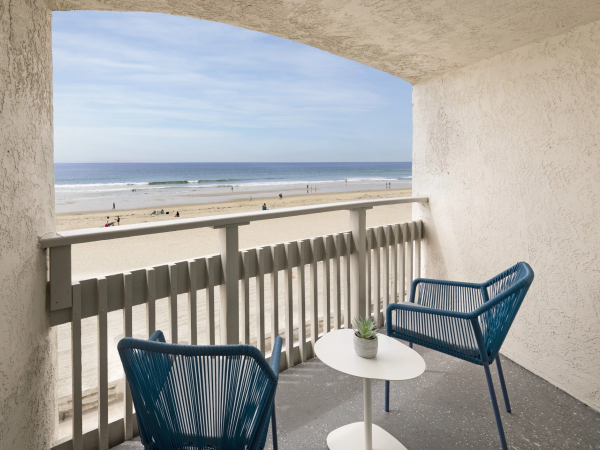 A balcony overlooking a sunny beach with two blue chairs and a small white table, inviting a relaxing sea view and gentle breeze.