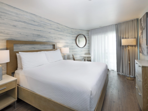 A modern hotel room with a large bed, nightstands, a round wall mirror, a desk, and a floor lamp, all in light wood tones and soft curtains.
