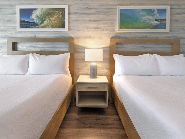 Two tidy hotel beds with white linens flank a shared nightstand and lamp, framed ocean pictures above on a light wood wall.