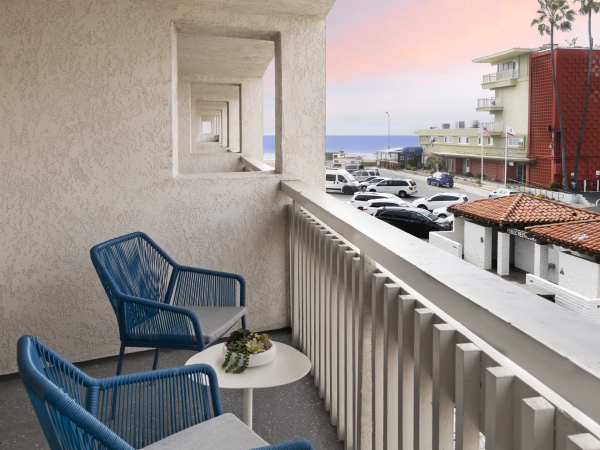 Two blue string chairs with a small round table on a balcony overlook a street and parked cars near the coast at sunset.