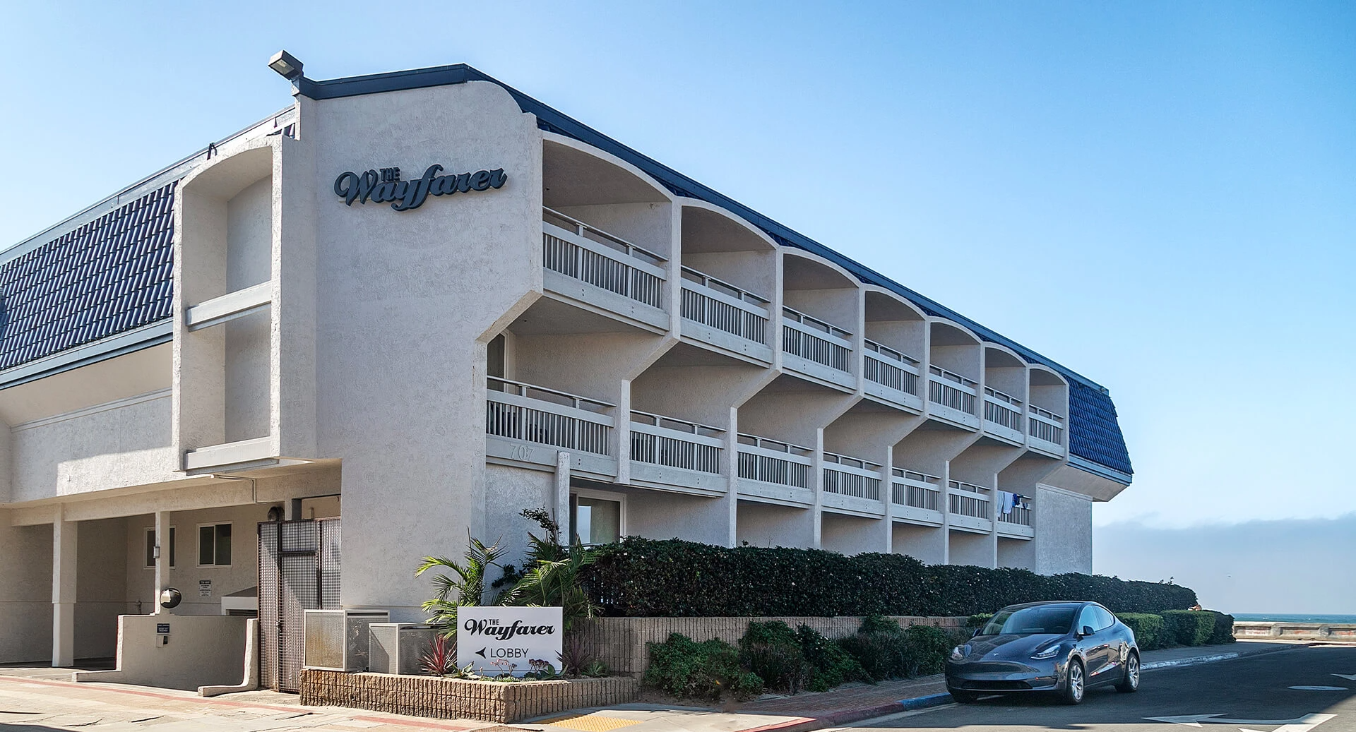 A white, multi-story motel with blue accents by the beach; balconies line the rooms, a parked car in front, and a &ldquo;Wharfstone&rdquo; sign near the entrance.