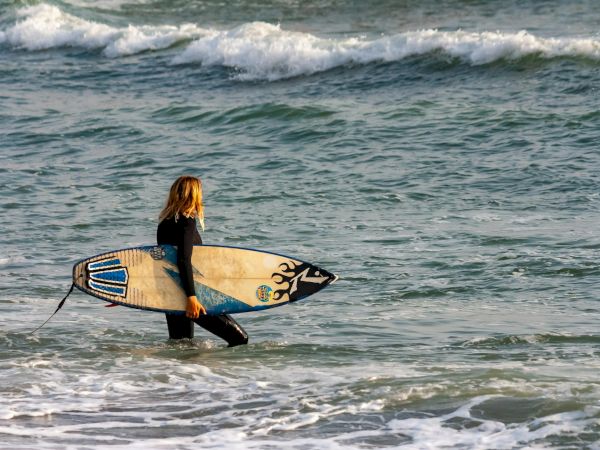 A person in a wetsuit carrying a surfboard along shallow ocean water, heading toward the waves.
