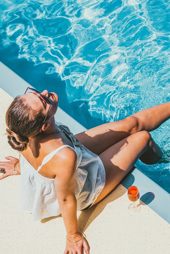 A woman sunbathes by a pool, legs in the water, towel on the edge, wearing a white swimsuit top; a bottle rests nearby, water glistening turquoise.