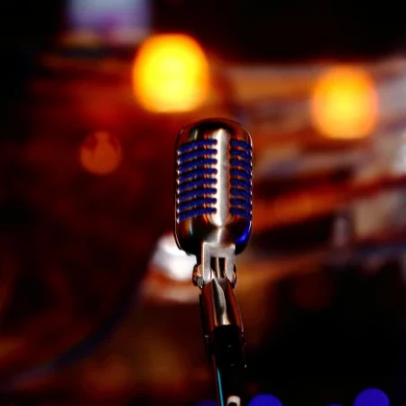 A close-up of a vintage microphone on a stand, spotlighted in a warm, blurred stage setting with colorful bokeh lights in the background.