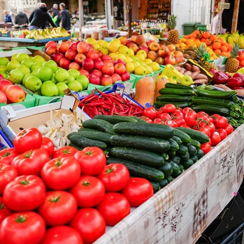 Colorful market stalls with fresh produce: tomatoes, peppers, cucumbers, apples, and other fruits arranged in crates and baskets along a busy outdoor street.