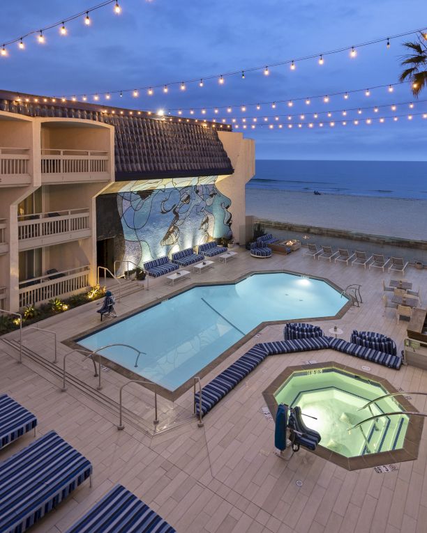 A tropical resort pool area at dusk with two pools, lounge chairs, string lights, palm trees, and the ocean in the background.