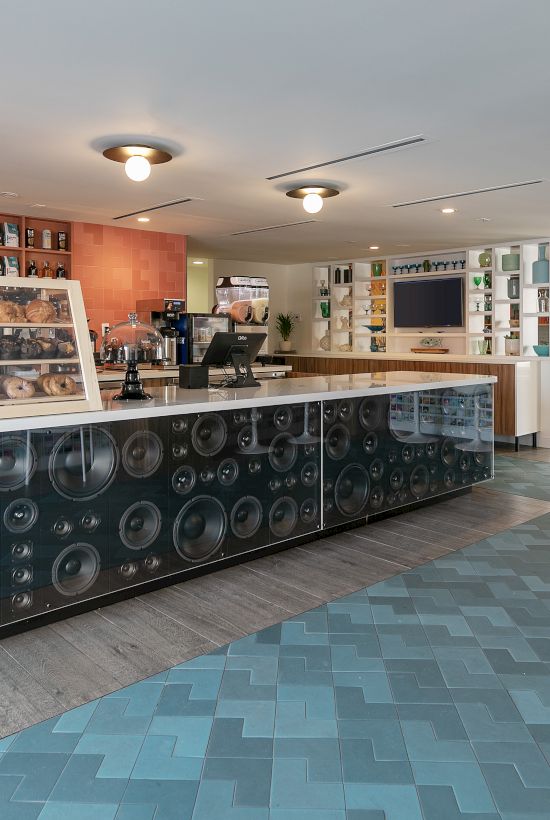 A retro-style cafe or shop counter with a long display case, beverage fridge, shelves, and tiled floor in a bright, airy space. End.