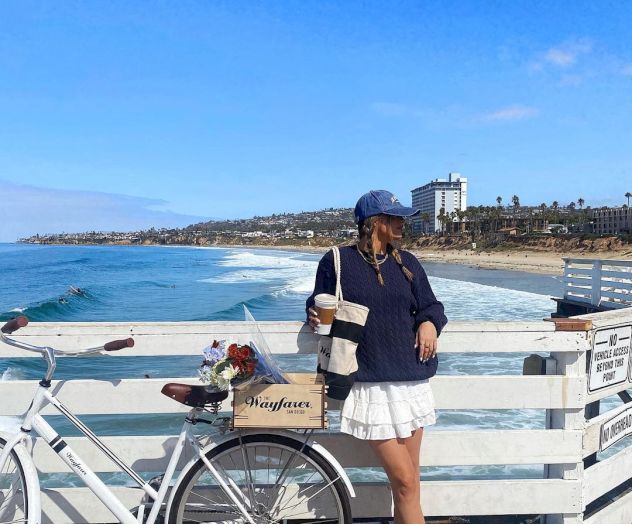 A woman in a navy top and white skirt stands on a pier with a white bicycle, blue sky, and ocean in the background, posing by the railing.