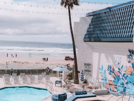 A sunny poolside scene with string lights, a tall palm, and a white building by the beach, blue accents, and people in the distance.