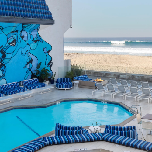 A bright poolside with blue lounge chairs, a mural of faces on the wall, and the beach and ocean beyond, under a clear sky, at a sunny resort.