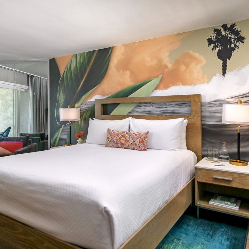 A modern hotel room with a neatly made bed, wall art, a desk, a chair, a flat-screen TV, and cozy warm lighting.