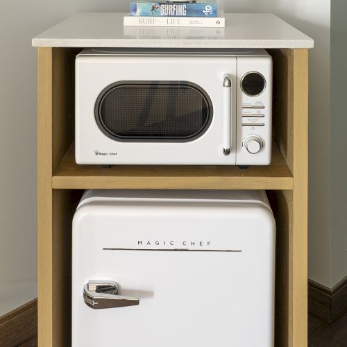 A tiny kitchen setup with a microwave on top of a small fridge, stacked on a wooden shelf, against a pale wall.