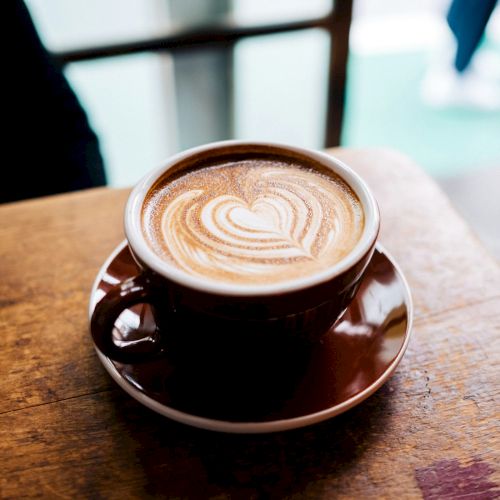 A close-up of a coffee cup on a saucer atop a wooden table, with steam rising and a blurred background.