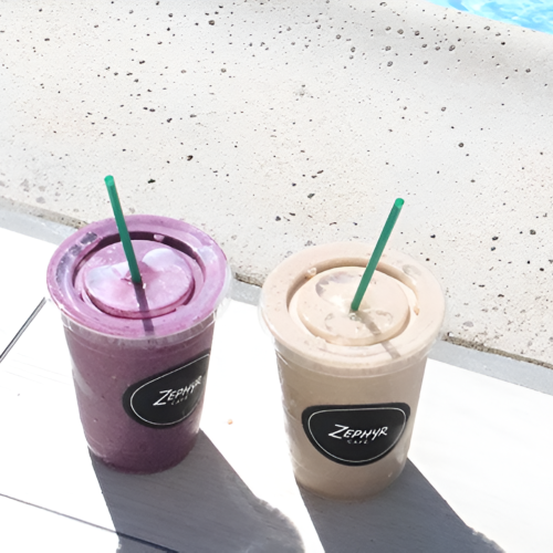 Two smoothie cups with green straws sit on a sunlit poolside ledge, one purple and one beige, with a pool edge in the background.