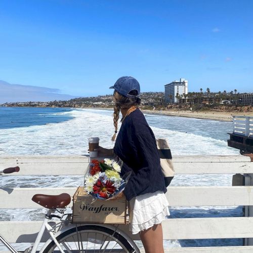 A girl in a white skirt and navy top with a blue cap stands by a pier, pushing a bicycle loaded with a basket of flowers toward the ocean, sunny day.