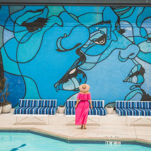 A woman in a pink dress stands by a pool with a blue mural of abstract faces behind her, by the poolside with striped loungers.