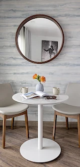 A small white round table with a vase of flowers, two beige chairs, a round wall mirror, and a framed black-and-white photo on marble wall.
