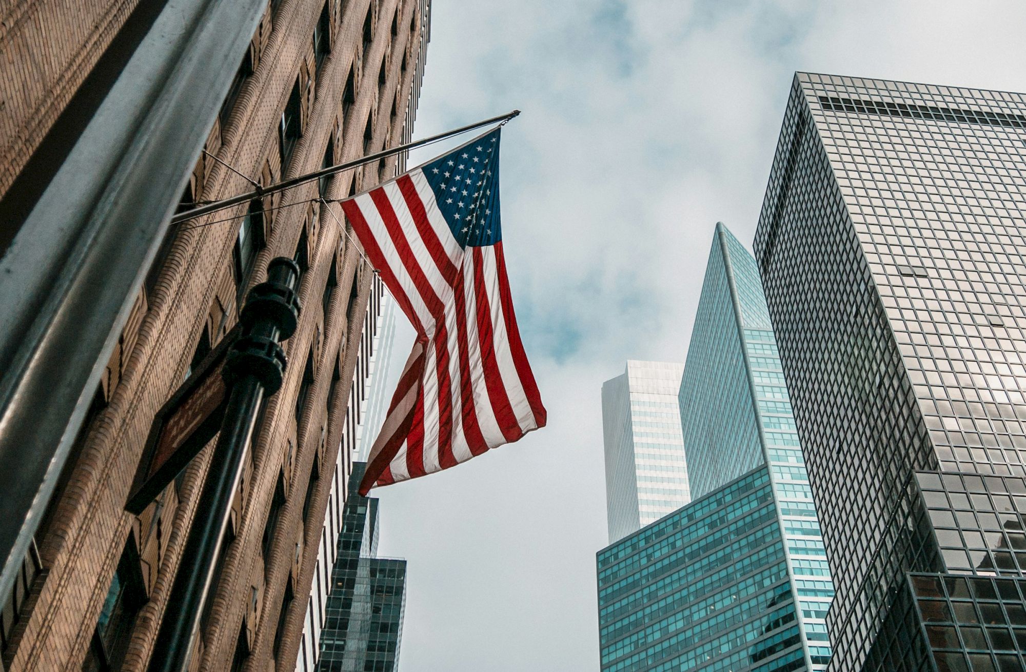 An American flag hangs from a tall building, with modern glass skyscrapers rising nearby against a cloudy sky.