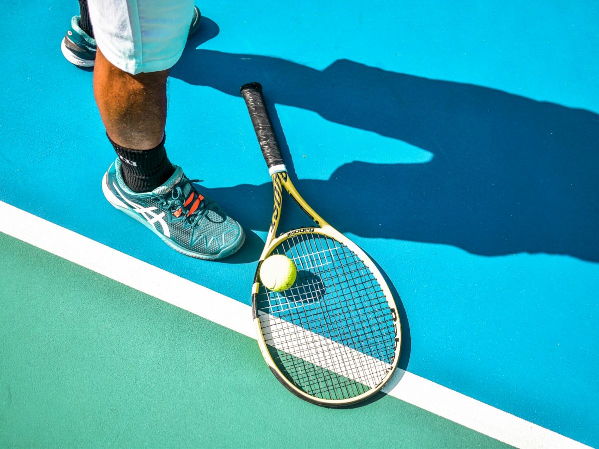 A tennis racket rests on a blue, green court beside a ball, with a player's shoe and shadow nearby, ready for a serve.
