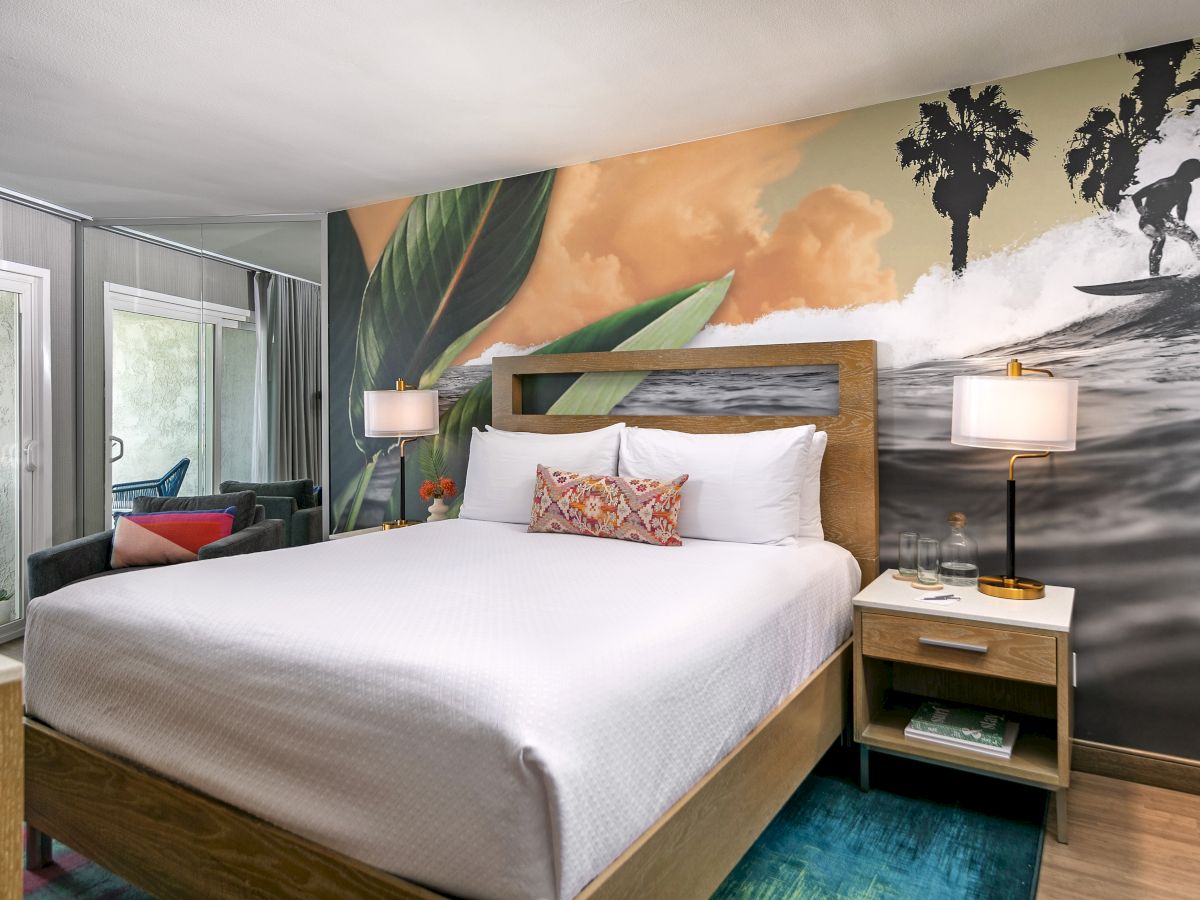A bright hotel room with a large bed, tropical mural wall, two nightstands, lamps, and a seating area by glass doors, inviting and stylish.